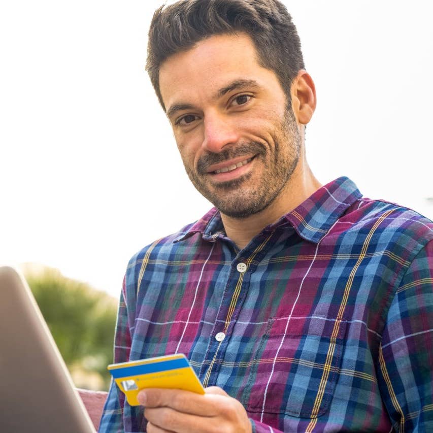 A man makes purchases with a credit card on his laptop in a city park.