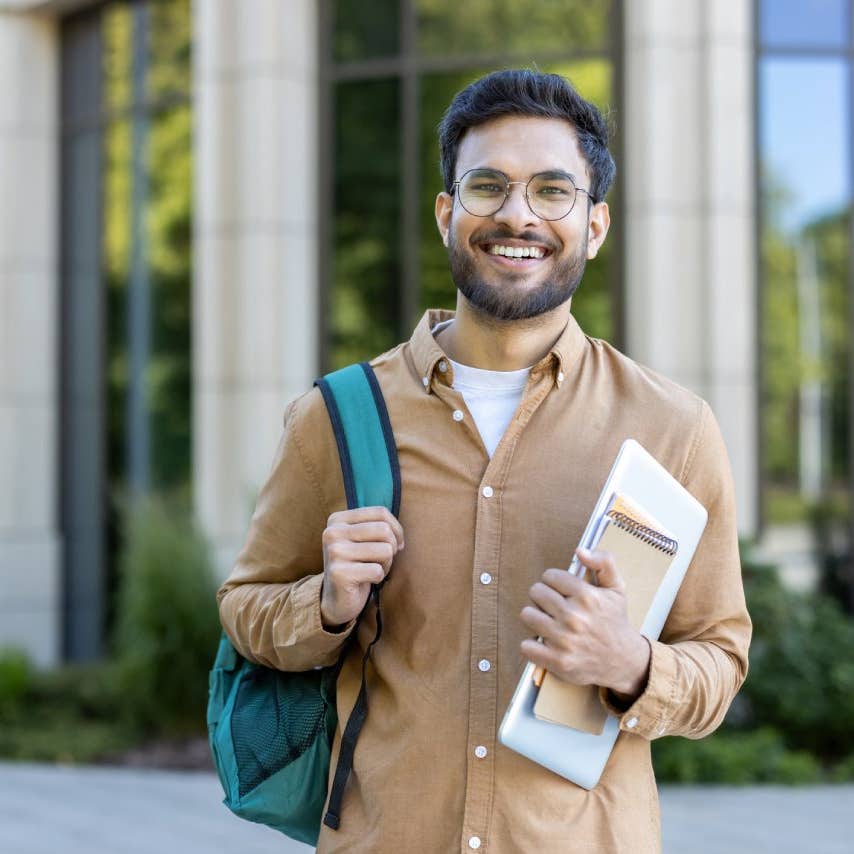 Smiling hispanic student stands outside academic building holding backpack and notebooks. Concept of higher education, confidence, and youth. Ideal for educational and lifestyle themes.