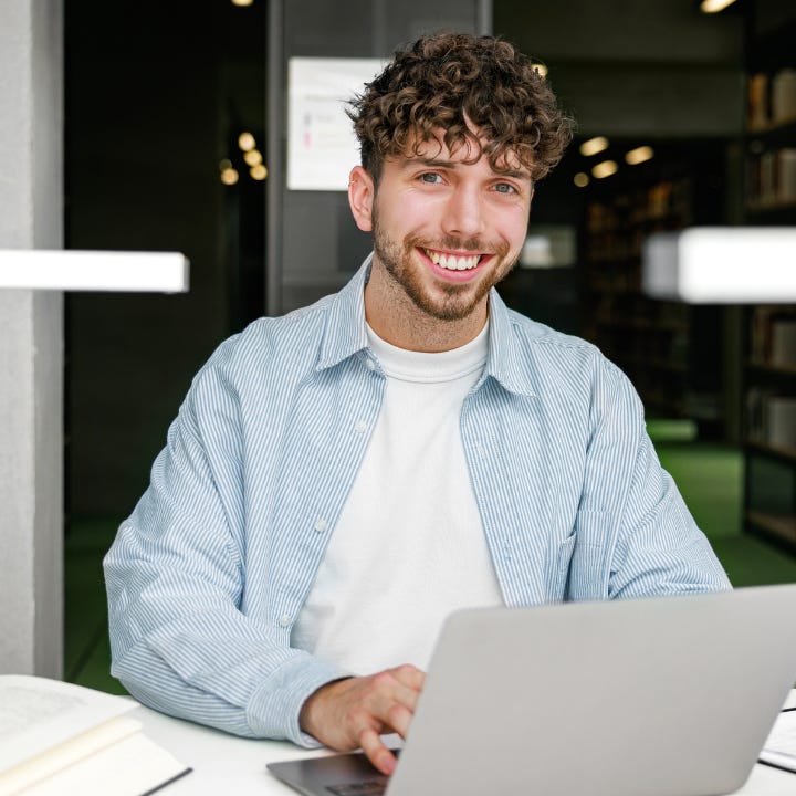 A portrait of a smiling young man works on his laptop with open books and notes nearby in a library. The inviting atmosphere and organized shelves emphasize focus and positivity in learning.